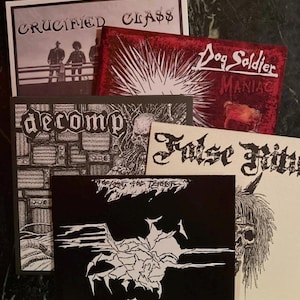May include: A collection of album covers with dark, gothic themes. The covers feature band names like "Crucified Class" and "Dog Soldier Maniac" in bold fonts. The artwork includes illustrations of figures, skulls, and abstract designs, with a predominantly black, white, and red color scheme.