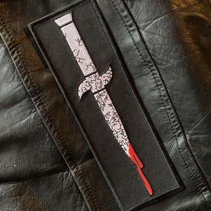 May include: A black patch with a white embroidered dagger with a red drip of blood. The dagger is stylized with a jagged edge and a curved handle.