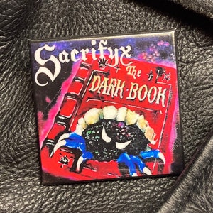Puede incluir: Un libro rojo con el título "The Dark Book" y el nombre de la banda "Sacrifyx" en una fuente gótica. El libro está abierto y una mano negra con garras azules sale de las páginas.