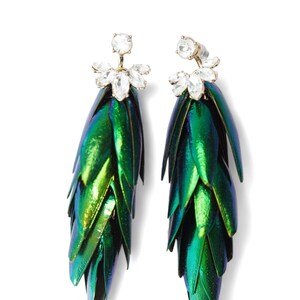 Op de afbeelding: Een paar bungelende oorbellen met iriserende groene kevervleugels. Elke oorbel heeft een cluster van heldere kristallen aan de bovenkant en een goudkleurige pin. De oorbellen zijn geplaatst tegen een witte achtergrond.