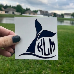 May include: A white sticker with a black whale tail and the letters "KLM" inside a circle. The sticker is held in a hand against a green grassy background.