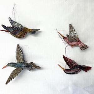 May include: Four colorful hummingbird ornaments with outstretched wings are displayed against a white background. The birds feature detailed patterns in shades of blue, brown, red, and silver, with delicate wire hangers.