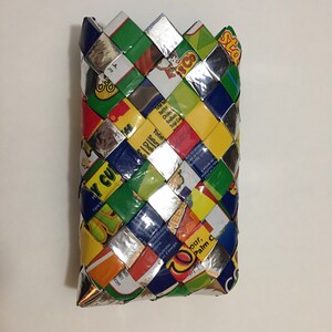 May include: A rectangular woven pouch made from colorful recycled packaging materials. The pouch features a variety of colors including green, yellow, blue, and silver. The woven design creates a textured surface.