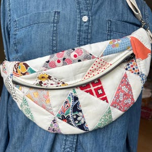 May include: A crescent-shaped crossbody bag constructed from quilted fabric featuring a patchwork of various colorful floral, geometric, and polka dot patterns in shades of red, blue, yellow, and white, secured with a black zipper closure.
