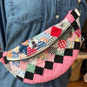 May include: A crescent-shaped fanny pack constructed from a patchwork quilt featuring a variety of colorful fabric squares in red, pink, black, blue, and white patterns. The bag includes a silver metal zipper closure and an adjustable white strap.