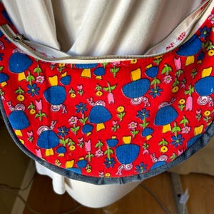 May include: A red corduroy fabric bag with a blue denim trim. The bag has a zipper closure and a whimsical pattern of blue mushrooms and yellow flowers on a red background.