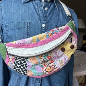 May include: A crescent-shaped fanny pack made from quilted patchwork fabric featuring various floral, polka dot, and geometric patterns in pink, yellow, green, and black. The bag includes a white zipper closure and an adjustable off-white strap.
