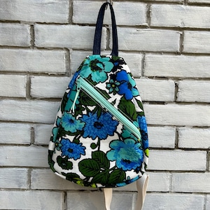 May include: A white, blue, and green floral print sling bag with a light green zipper. The bag has a black strap and a white strap.