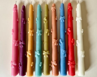 May include: Nine brightly coloured taper candles. The candles are in shades of pink, purple, orange, teal, olive green, peach, blue, red, and white. Each candle is decorated with small bow accents.