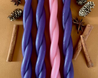 May include: Four twisted candles, two purple and two pink, arranged in a row. The candles are on a brown surface with cinnamon sticks and pine cones.