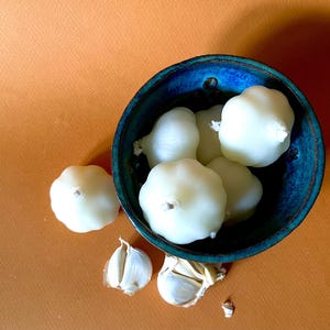 May include: A blue ceramic bowl filled with white, peeled garlic cloves. Two cloves and two garlic sections are next to the bowl on an orange surface. The garlic cloves have a round shape with a small stem.