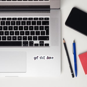 May include: A silver laptop computer with a black keyboard and a sticker that says "get shit done" on the bottom right corner of the trackpad. The laptop is on a white surface with a black cell phone, a black pencil, and a blue pen.