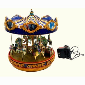 May include: A decorative carousel music box featuring a blue and gold canopy with ornate mirrors, rotating miniature horses and figures on a circular base, accompanied by a black electrical power adapter with a cord for operation.