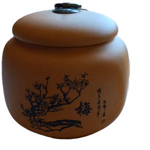 May include: A round, matte terracotta-colored ceramic jar with a matching lid. The lid features a dark, circular metal handle. The body of the jar is decorated with a black ink-style illustration of a flowering branch and black Chinese calligraphy.