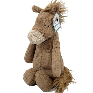 May include: A plush horse toy made of soft, light brown fur with a lighter beige muzzle. The stuffed animal features a shaggy mane and tail, and a white tag attached to the ear with the text Jellycat London and a black cat logo printed on it.