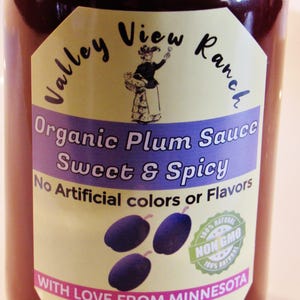 May include: A glass jar of Valley View Ranch Organic Plum Sauce, Sweet & Spicy. The label is yellow with a picture of a woman in a dress and a plum design. The label also says "No Artificial colors or Flavors" and "100% Natural Non GMO 100% Natural". The label also says "With Love From Minnesota" and "Net wt 9 oz (252g) Good Up To 2 Years".