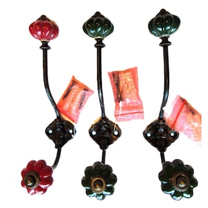 May include: Three wall-mounted coat hooks with black metal frames and decorative ceramic knobs. One hook features a red knob, while the other two have dark green knobs. Each hook includes a small plastic bag containing mounting hardware and screws.