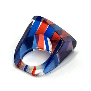 May include: A chunky ring crafted from translucent material featuring a geometric pattern of bold, diagonal stripes in vibrant shades of blue, red, and purple. The ring has a smooth, polished finish with a wide, angular band design.