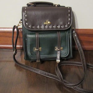 May include: A two-tone crossbody bag with a flap closure. The top flap is brown, and the main body is dark green. It features white stitching, gold-tone hardware, and a bee emblem. The bag has a long, adjustable strap.