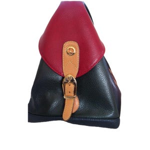 May include: A triangular-shaped backpack featuring a textured red leather flap over a dark navy blue body. A tan leather strap with a gold-toned metal buckle closure is centered on the front. The buckle hardware displays a small Eiffel Tower emblem.
