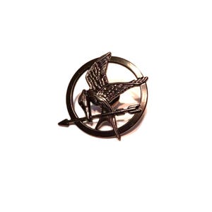 May include: A dark metal pin featuring a stylized bird with outstretched wings, perched atop an arrow. The bird and arrow are set within a circular frame. The pin has a metallic finish and is set against a white background.