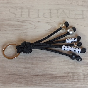 Könnte beinhalten: Schwarzer Paracord-Schlüsselanhänger mit einem silbernen Schlüsselring und mehreren Perlen. Die Perlen buchstabieren die Namen "Jerry" und "Tom".