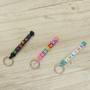 Könnte beinhalten: Drei personalisierte Schlüsselanhänger mit Namen, die in bunten Buchstabenperlen geschrieben sind. Jeder Schlüsselanhänger hat eine unterschiedlich farbige Kordel und einen silbernen Schlüsselring. Die Namen sind Stefan, Geena und Julius.