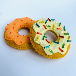 Op de afbeelding: Twee donuts van vilt, een met oranje sprinkles en een met witte glazuur en kleurrijke sprinkles.