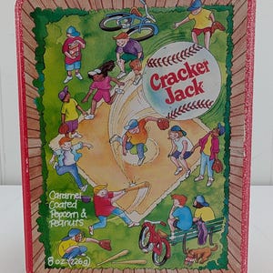 May include: Rectangular Cracker Jack tin with a baseball-themed illustration. The tin features a baseball, a baseball field, and children playing baseball. The text "Cracker Jack" is on the baseball. The tin also says "Caramel Coated Popcorn & Peanuts" and "8 oz (226g)."