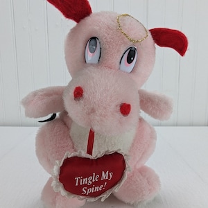 May include: A pink plush hippo toy with red ears and accents, a gold halo, and large eyes. It holds a red heart-shaped pillow with the text "Tingle My Spine!" in white. The toy is on a white surface.