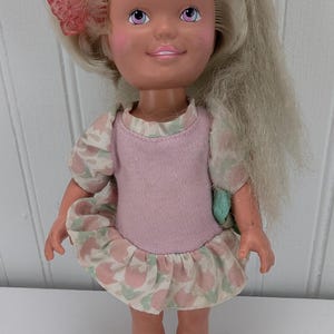 Puede incluir: Una muñeca de plástico vintage con cabello rubio, mejillas rosadas y ojos morados. Lleva un vestido floral rosa y verde y zapatos verdes.