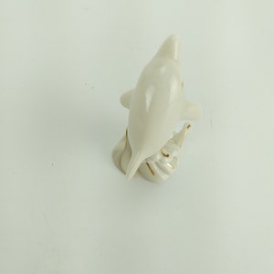 May include: White porcelain figurine of a dolphin with gold accents. The dolphin is leaping out of the water and has its tail raised.