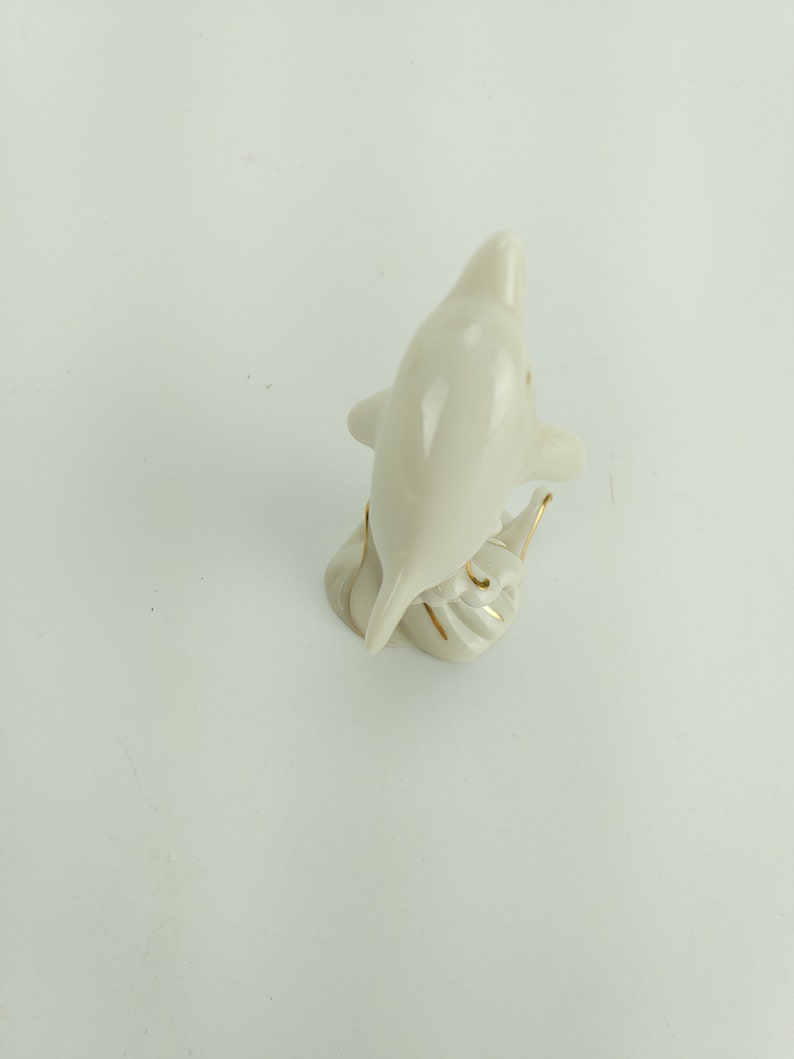 May include: White porcelain figurine of a dolphin with gold accents. The dolphin is leaping out of the water and has its tail raised.