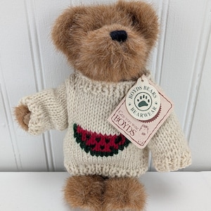 Puede incluir: Un oso de peluche marrón que lleva un suéter color crema con un diseño de sandía. El oso tiene una etiqueta que dice "Boyds Bears Bearwear". El pelaje del oso es de color marrón claro.