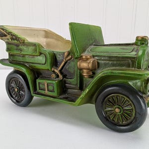 May include: A green vintage car planter with black wheels and gold accents. The planter has a detailed design, including a driver's seat and a compartment for plants. The car is made of a durable material and has a distressed finish.