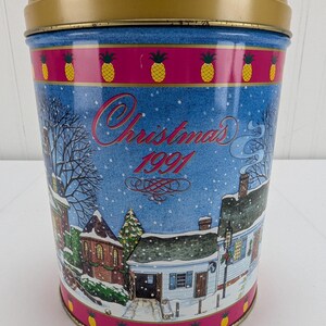Könnte beinhalten: Eine blaue und weiße Weihnachtsdose mit einer Winterszene und dem Text "Christmas 1991". Die Dose hat einen goldenen Deckel und ein rosafarbenes Band mit gelben Ananas.