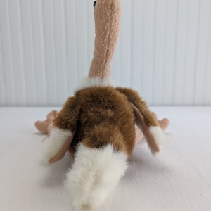 Puede incluir: Un juguete de peluche de avestruz marrón y blanco con un cuello largo y una cola blanca. El juguete está acostado de espaldas con la cabeza mirando hacia la cámara.