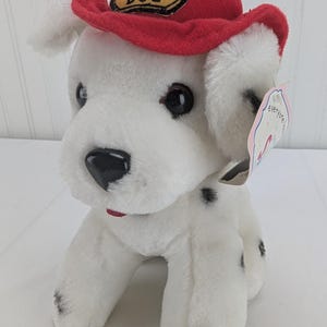 May include: A plush white Dalmatian puppy toy with black spots, wearing a red felt firefighter hat featuring a yellow patch that reads FIRE DOG. The stuffed animal has large dark plastic eyes, a black nose, and a small paper tag attached.