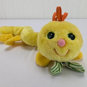 May include: A plush yellow caterpillar toy with a smiling face, black eyes, and a pink nose. It has orange antennae and a green and white striped scarf. The caterpillar's body is segmented, with yellow feet.