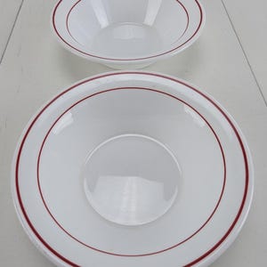 Puede incluir: Dos cuencos blancos con borde rojo. Los cuencos son de diferentes tamaños, con el cuenco más pequeño en la parte superior. Los cuencos están hechos de un material brillante y están colocados sobre un fondo blanco. Los cuencos son probablemente para servir comida.