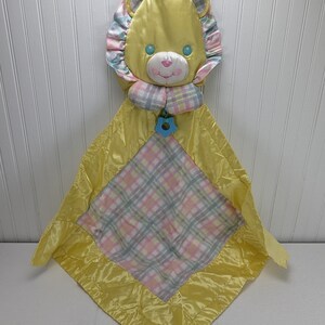 May include: A yellow satin baby blanket with a plaid center and a lion head attached. The lion has a blue and pink plaid mane and blue eyes.