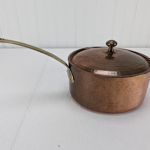 May include: A copper saucepan with a lid and a long brass handle. The pot has a hammered texture and a brass knob on the lid. The handle is attached to the side of the pot with a brass rivet. The saucepan is designed for cooking.