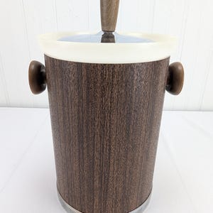 May include: A vintage ice bucket with a dark brown wood grain pattern. It has a white rim, a clear lid, and wooden handles. The lid has a wooden handle. The base is trimmed with a silver band.