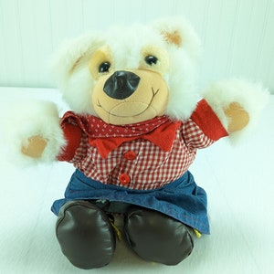 Puede incluir: Oso de peluche blanco con una camisa de cuadros rojos y blancos, una falda vaquera azul y botas marrones. El oso tiene una nariz negra y ojos marrones.