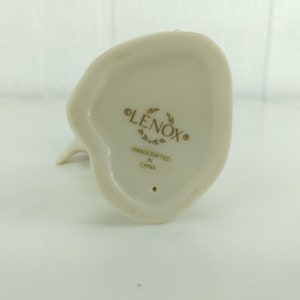 May include: The bottom of a white porcelain figurine with the words "LENOX" and "HANDCRAFTED IN CHINA" in gold lettering.