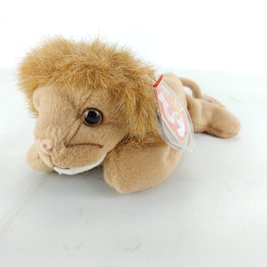 May include: A soft toy lion in shades of brown and tan, with a fluffy mane and a small tag. The lion is lying down, with its head and front paws visible. The toy is made of a soft material.