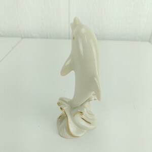 May include: A white porcelain dolphin figurine with gold accents, leaping out of the water. The dolphin is positioned vertically, with its tail raised and its head tilted back.