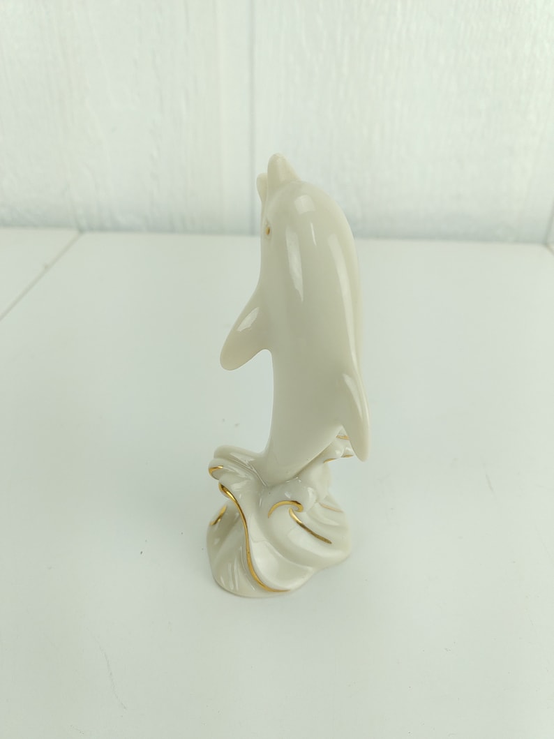 May include: A white porcelain dolphin figurine with gold accents, leaping out of the water. The dolphin is positioned vertically, with its tail raised and its head tilted back.