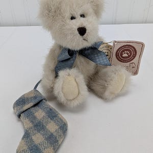 Pode incluir: Um urso de pelúcia creme com uma fita azul e uma pequena meia xadrez combinando. O urso tem um nariz marrom escuro e olhos pretos pequenos. Uma etiqueta está presa ao urso.
