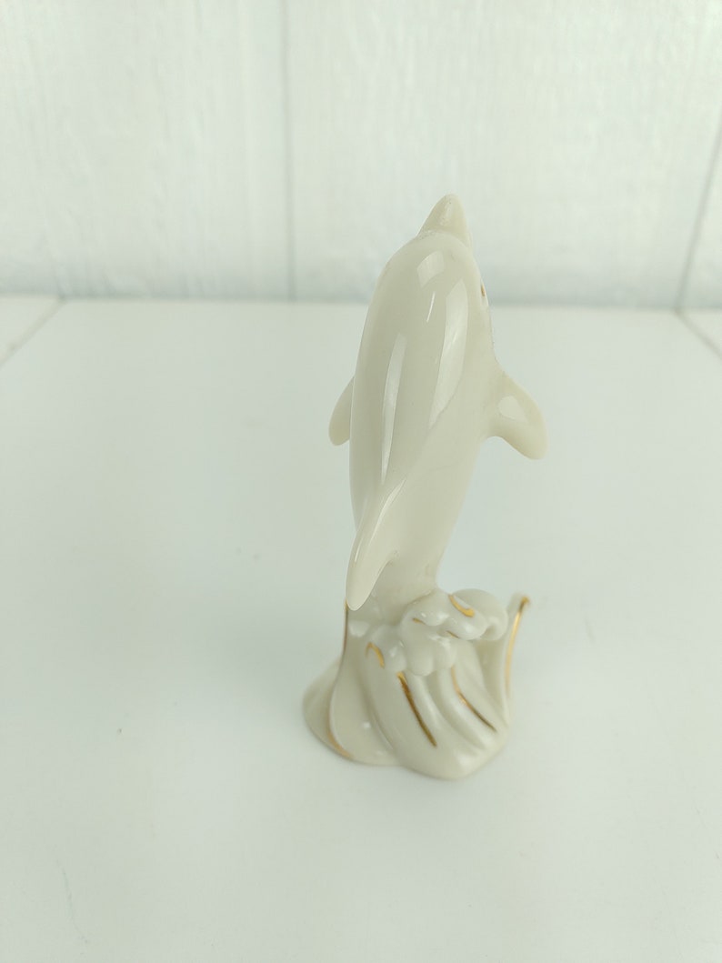 May include: A white porcelain figurine of a dolphin leaping out of the water. The dolphin is detailed with gold accents and is set on a base with a wave design.